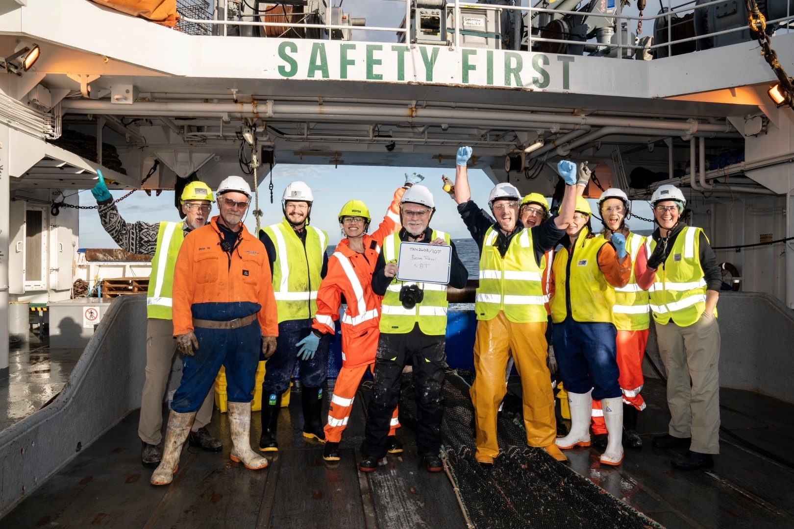 Smiling group of researchers wearing safety gear posing for a photograph on the stern of RV Tangaroa.