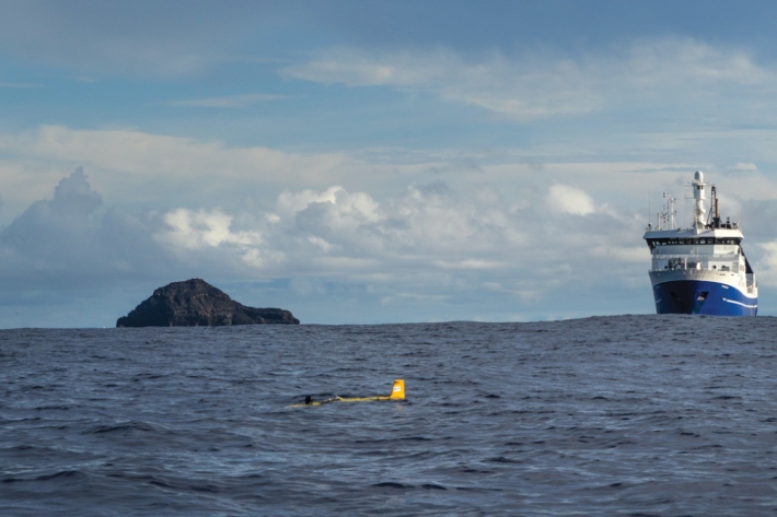 A remote ocean glider gathers oceanographic readings as RV Tangaroa sits on station with the blackened remnants of Hunga Tonga-Hunga Ha'apai volcano to starboard.