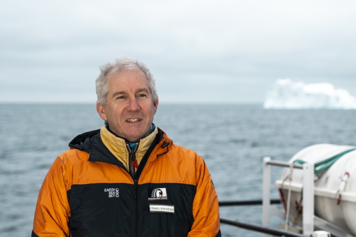 Close up shot of Craig Stevens, standing against a ship railing with view of Antarctic Ocean. An iceberg is seen on the right side.