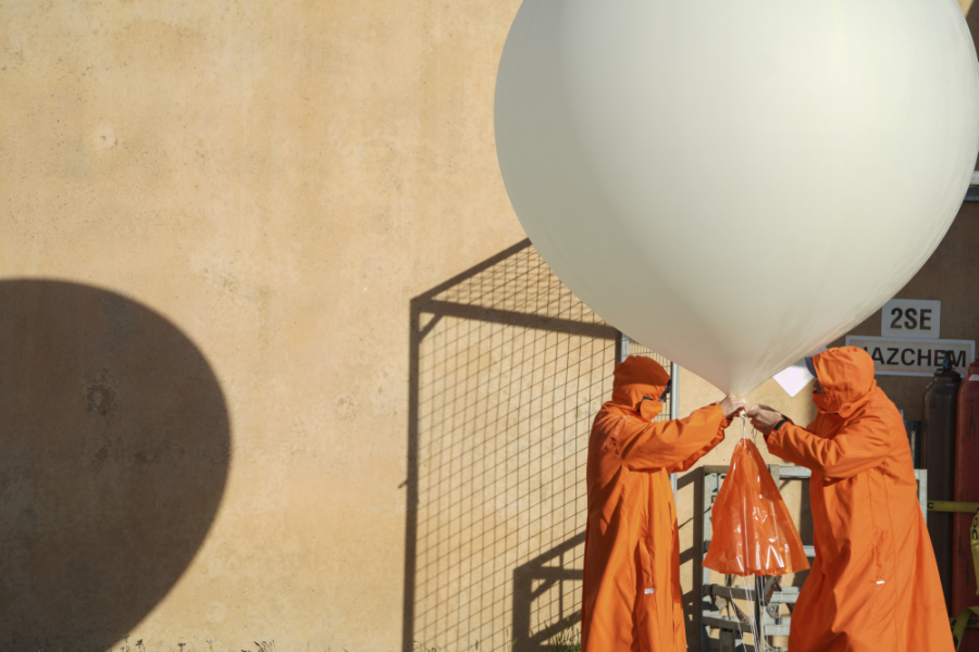 Two scientists clad in orange coats holding up inflated white balloon which is attached to an orange plastic bag which has the ozonesonde instrument.