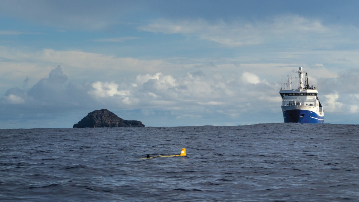 A remote ocean glider gathers oceanographic readings as RV Tangaroa sits on station with the blackened remnants of Hunga Tonga-Hunga Ha'apai volcano to starboard.