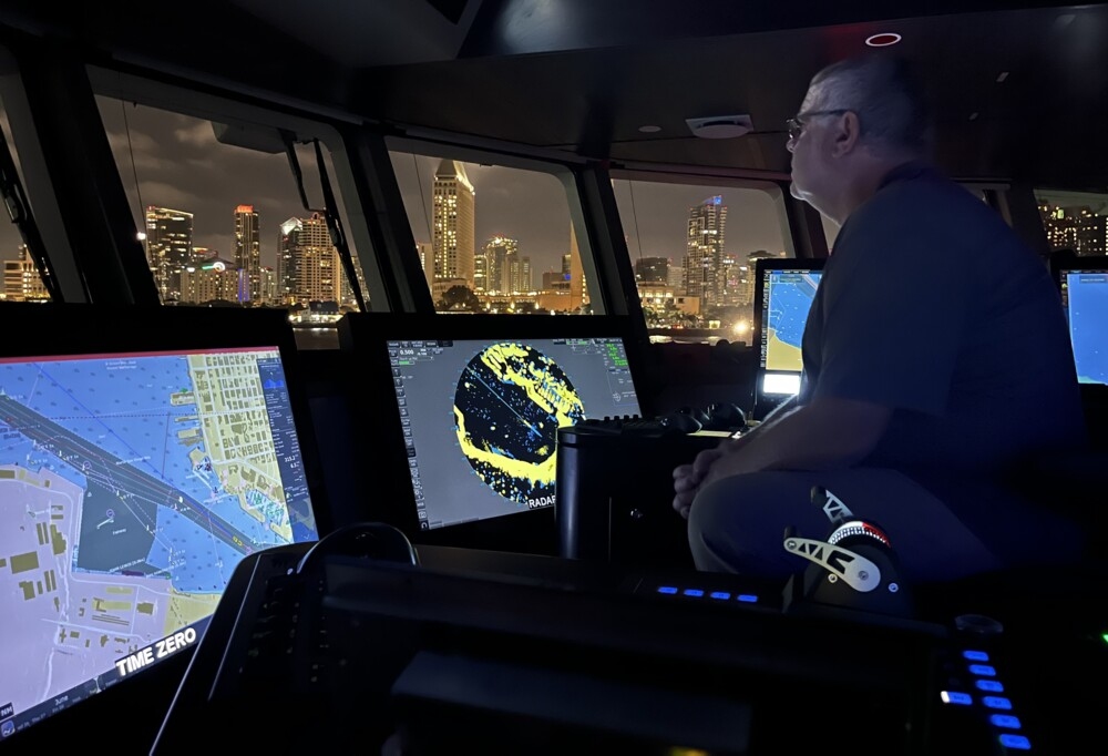Chris Whalan, Captain of Kaharoa II, navigates the ship in San Diego harbour. The cityscape in the background is so close it seems as though he is driving up the street.